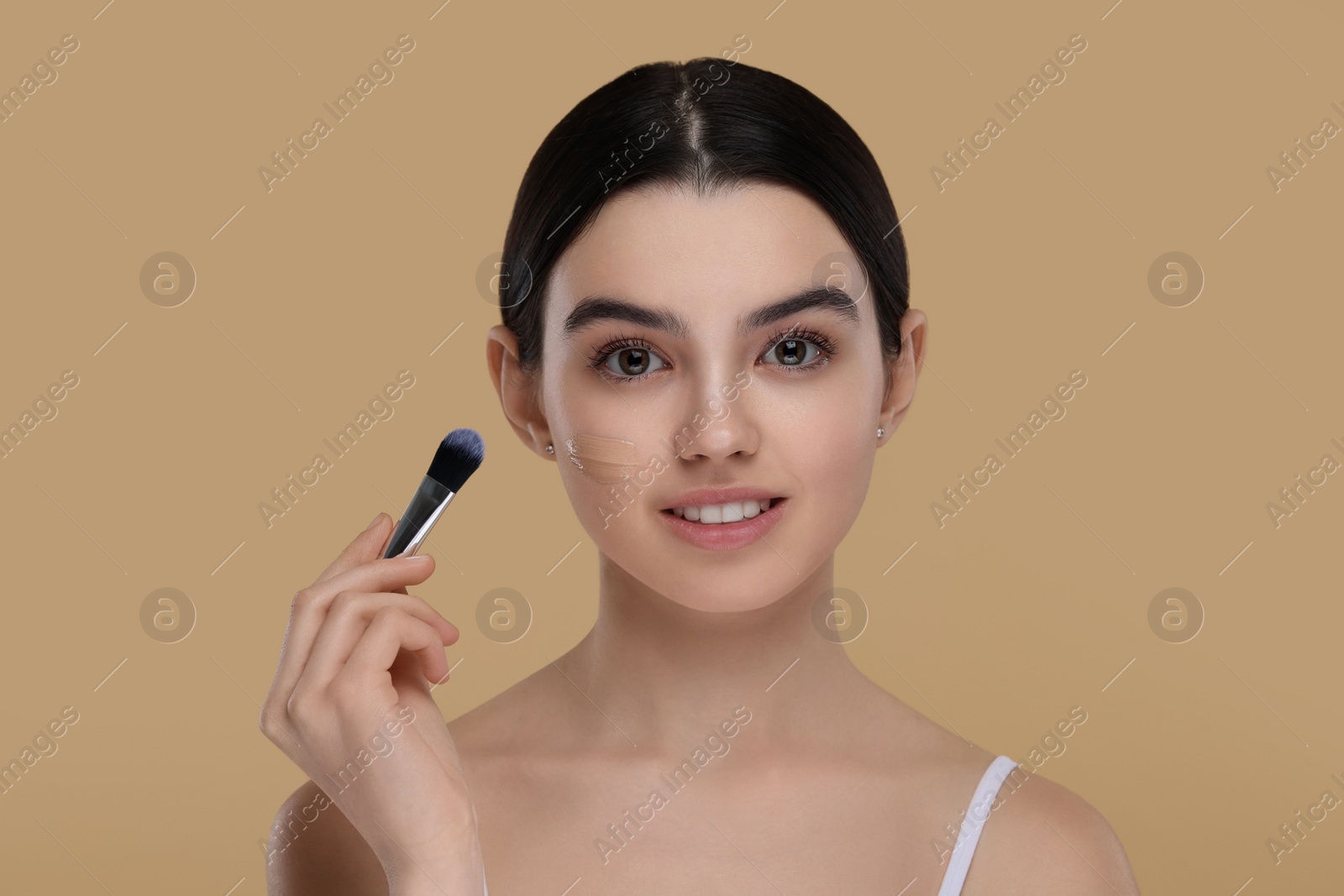 Photo of Teenage girl applying foundation on face with brush against beige background