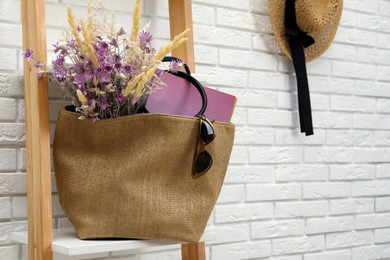 Stylish beach bag with beautiful bouquet of wildflowers, sunglasses and magazines on shelf indoors. Space for text