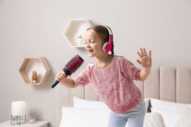Photo of Cute little girl in headphones with hairbrush singing on bed at home