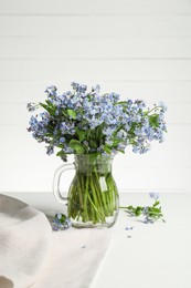 Photo of Bouquet of beautiful forget-me-not flowers in glass jug and blue cloth on white table