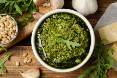 Photo of Bowl of tasty arugula pesto and ingredients on wooden table, flat lay