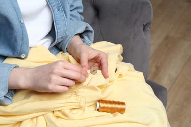 Photo of Woman sewing button with needle and thread onto shirt at home, closeup