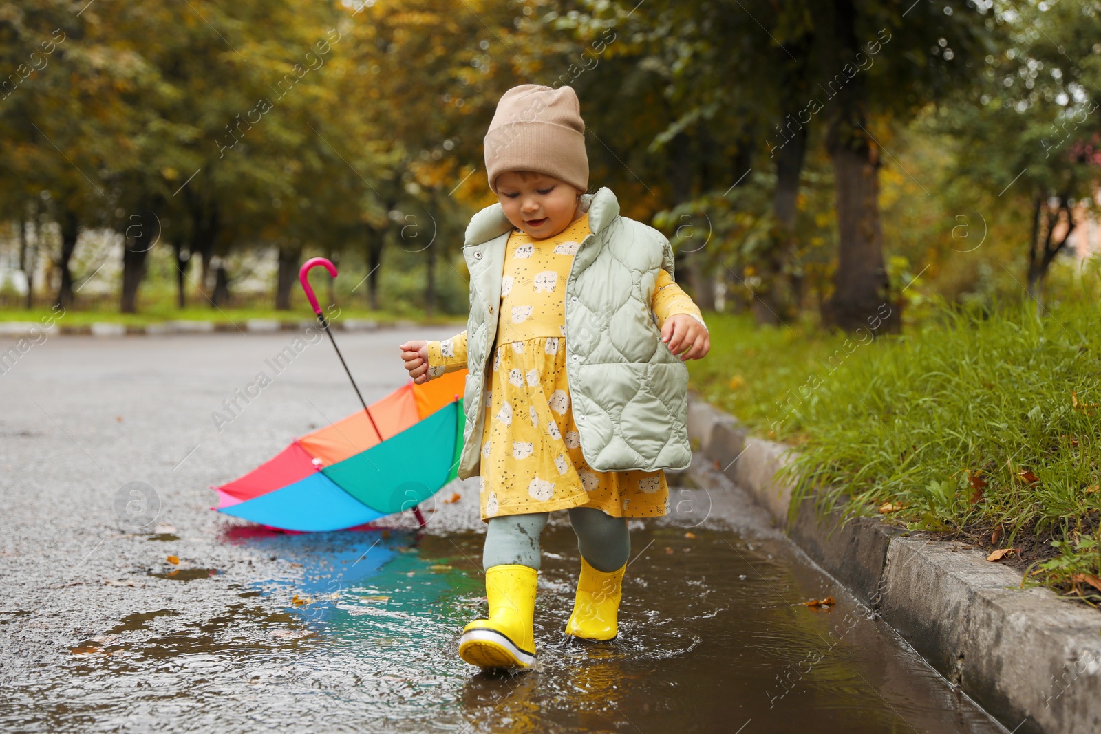 Photo of Cute little girl splashing water with her boots in puddle outdoors