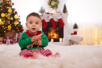 Baby wearing cute elf costume on floor in room decorated for Christmas