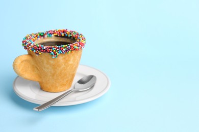 Photo of Delicious edible biscuit coffee cup decorated with sprinkles and spoon on light blue background, space for text