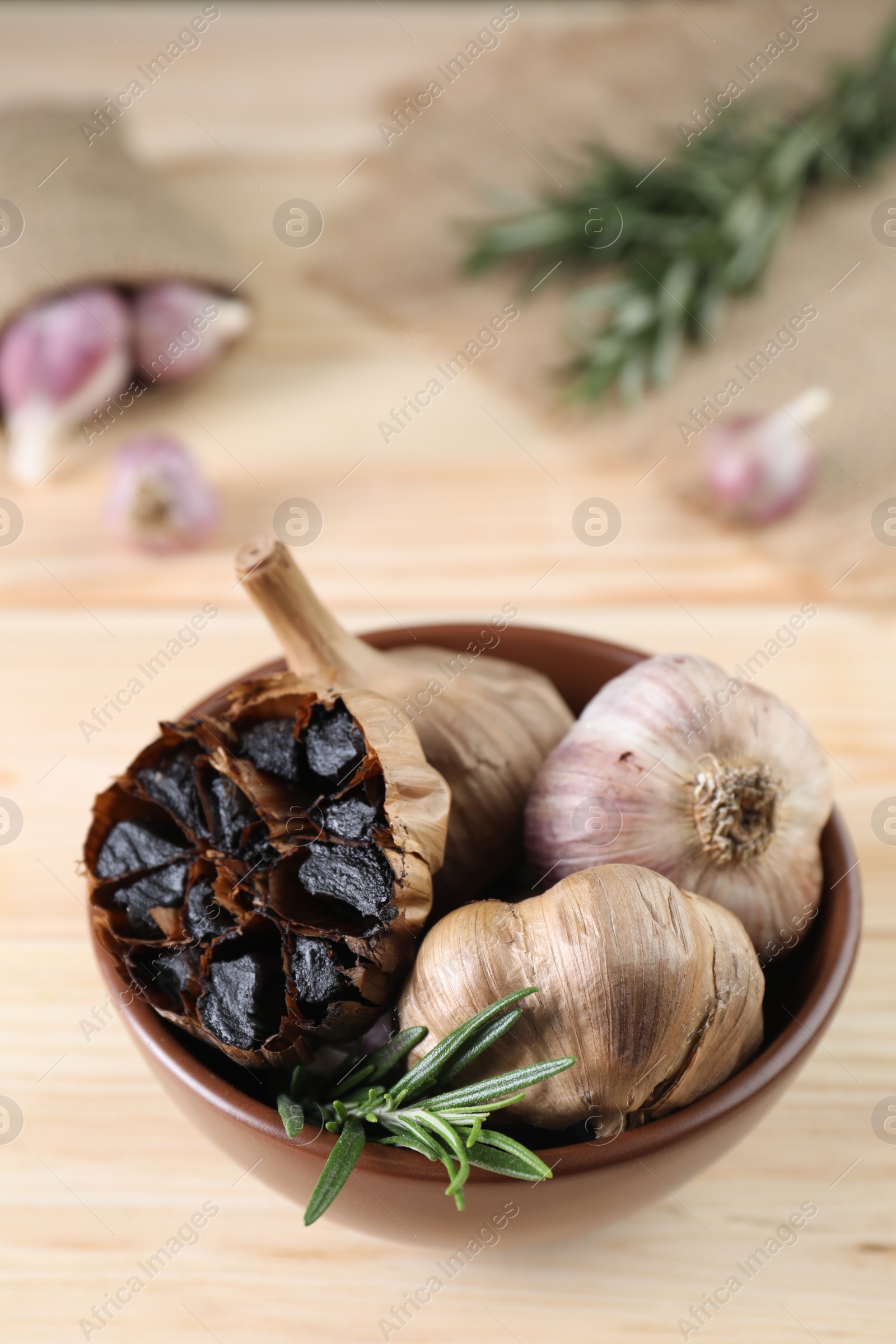 Photo of Bulbs of fermented black garlic and rosemary in bowl on wooden table