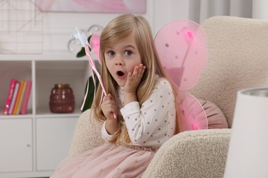 Photo of Surprised little girl in fairy costume with pink wings and magic wand in armchair at home