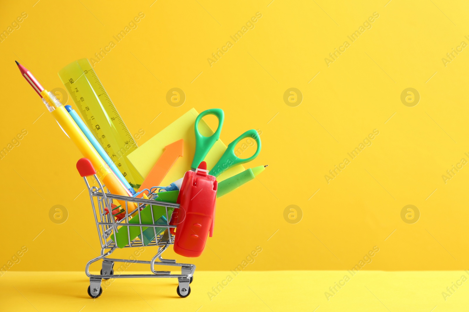 Photo of Small shopping cart with different school stationery on yellow background. Space for text