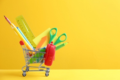 Photo of Small shopping cart with different school stationery on yellow background. Space for text