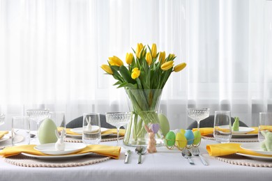 Photo of Festive table setting with glasses, painted eggs and vase of tulips. Easter celebration