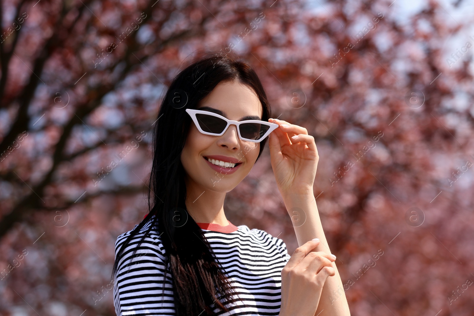 Photo of Pretty young woman with sunglasses near beautiful blossoming trees outdoors. Stylish spring look