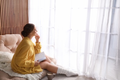 Beautiful young woman with book and cup of coffee near window at home, space for text. Winter atmosphere