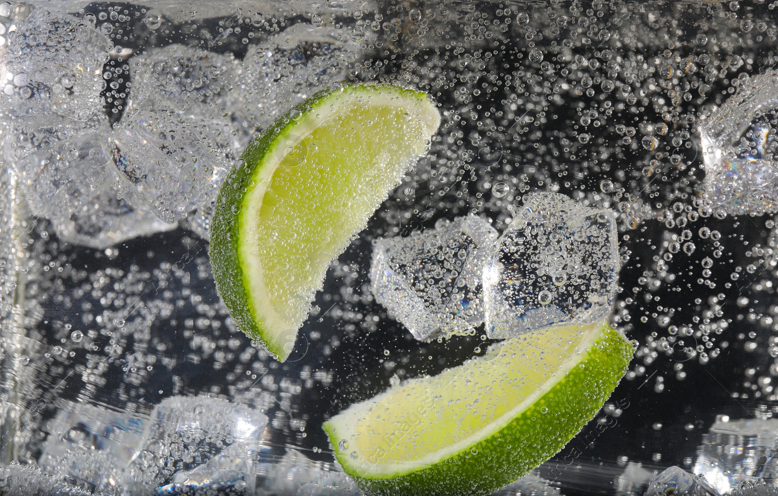 Photo of Juicy lime slices and ice cubes in soda water against black background, closeup