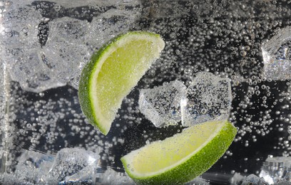 Photo of Juicy lime slices and ice cubes in soda water against black background, closeup