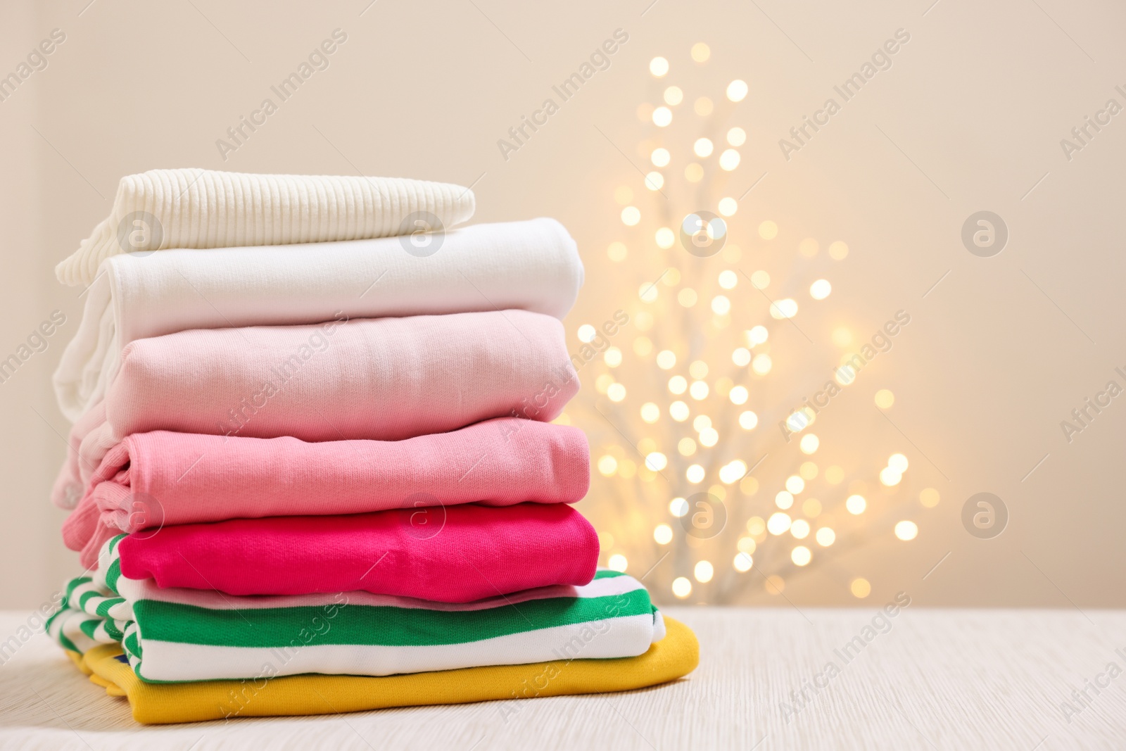 Stack of folded clothes on wooden table against beige background with blurred lights, space for text Photo of Stack of folded clothes on wooden table against beige background with blurred lights, space for text