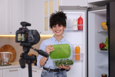 Smiling food blogger opening container with spinach while recording video in kitchen