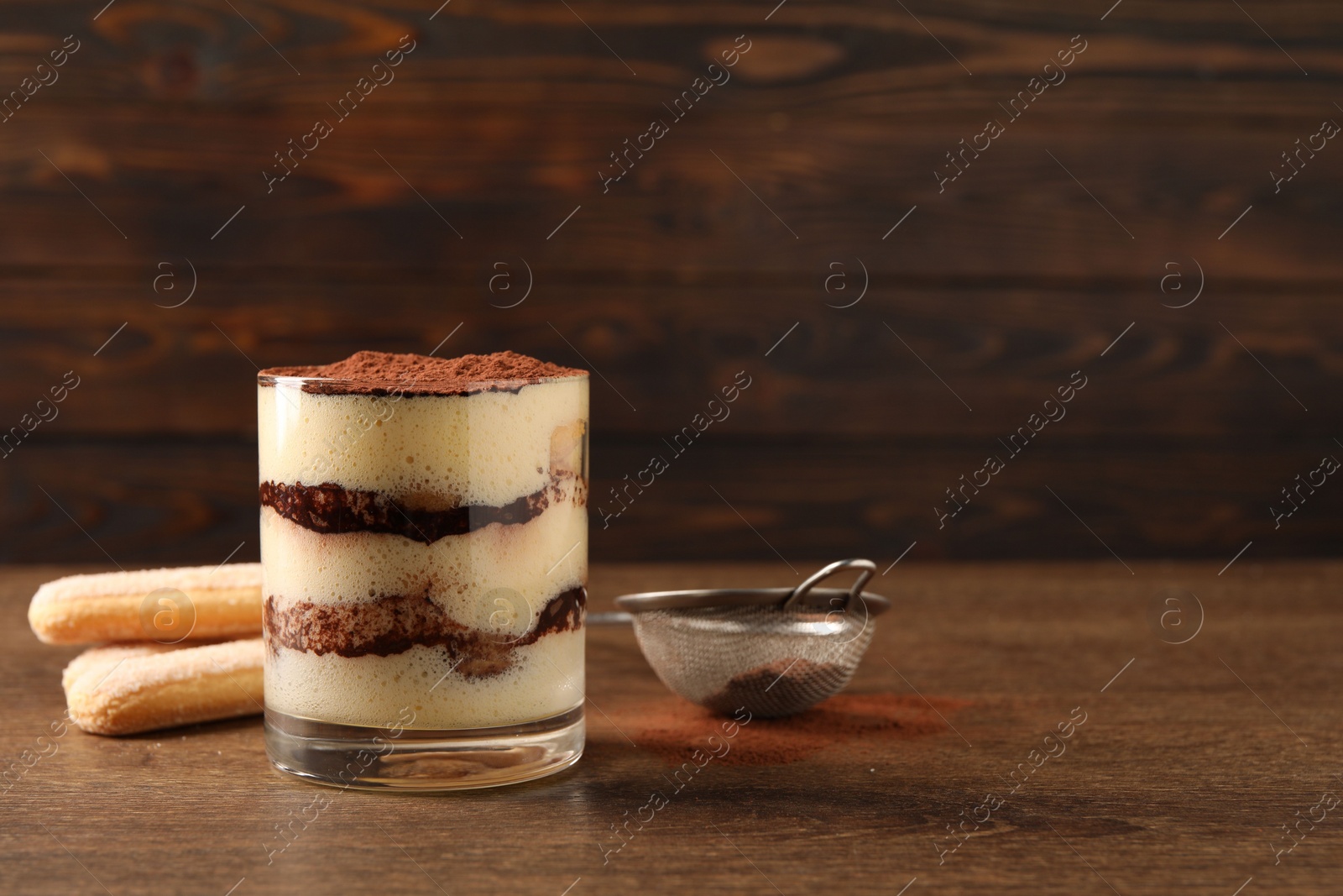 Photo of Delicious tiramisu in glass, biscuits and sieve with cocoa powder on wooden table. Space for text