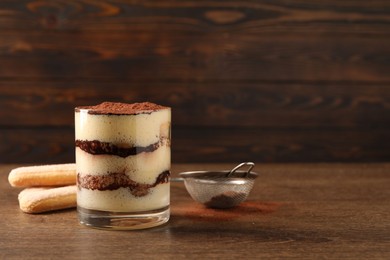 Photo of Delicious tiramisu in glass, biscuits and sieve with cocoa powder on wooden table. Space for text