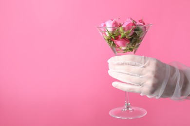 Woman in elegant gloves holding beautiful martini glass with water and roses on pink background, closeup. Space for text