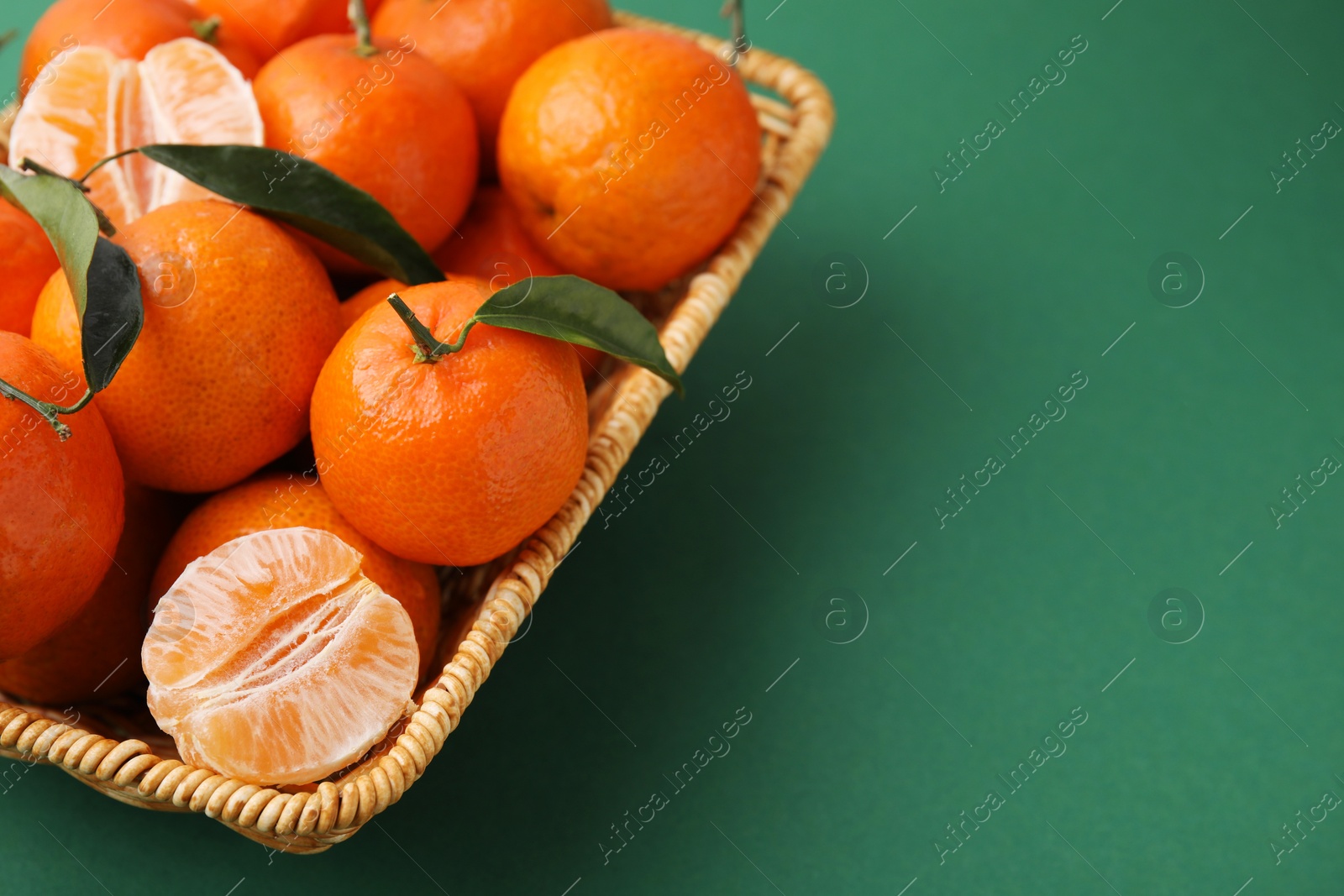 Photo of Fresh ripe tangerines with leaves in wicker basket on green background, closeup. Space for text