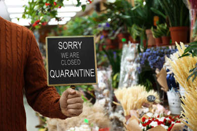 Business owner holding sign with text SORRY WE ARE CLOSED QUARANTINE in flower shop, closeup