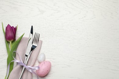Photo of Cutlery set, painted eggs and beautiful flower on white wooden table, flat lay with space for text. Easter celebration