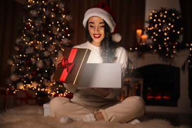 Photo of Young woman wearing Santa hat opening Christmas gift on floor at home