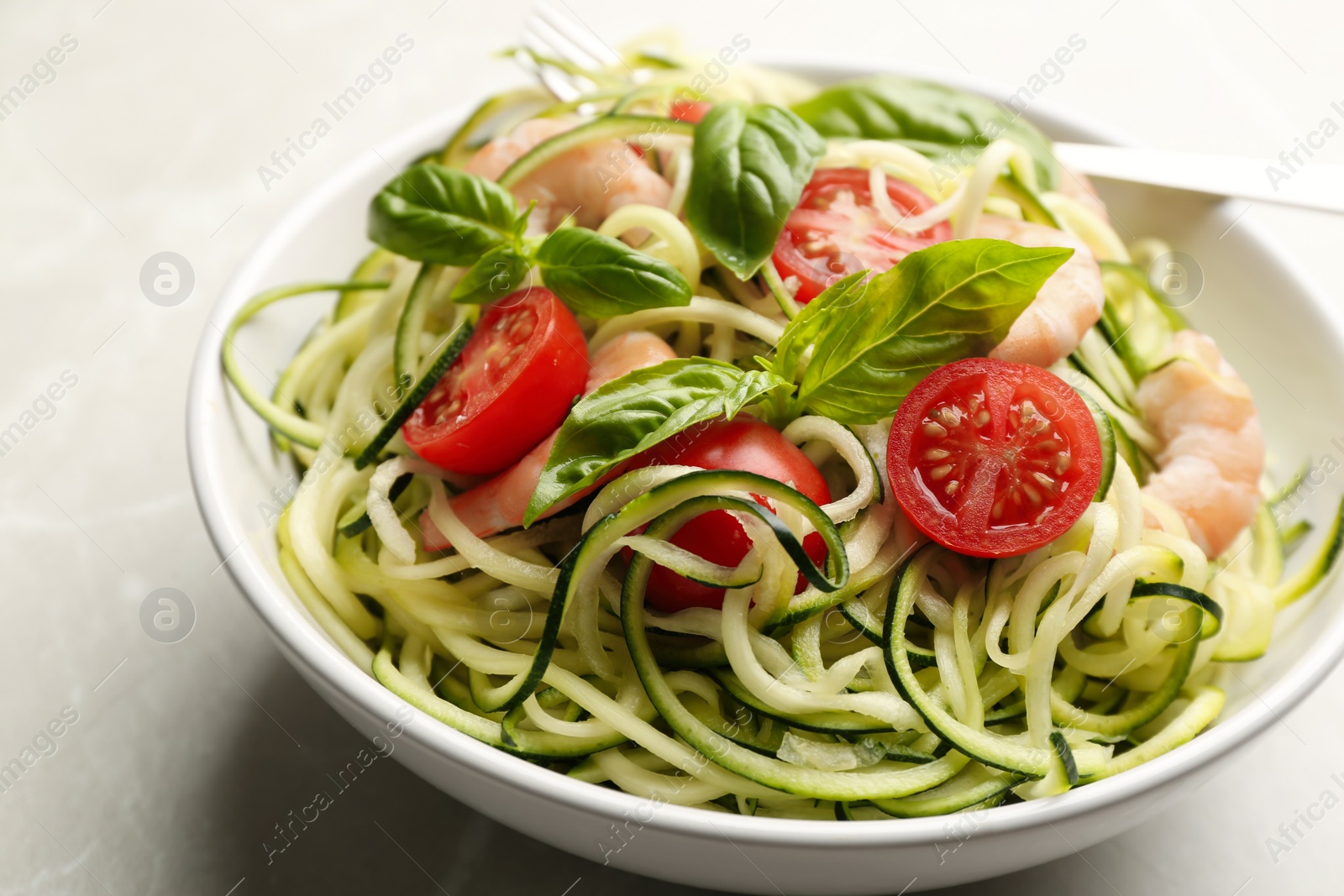 Photo of Delicious zucchini pasta with shrimps, cherry tomatoes  and basil in bowl on light grey table, closeup