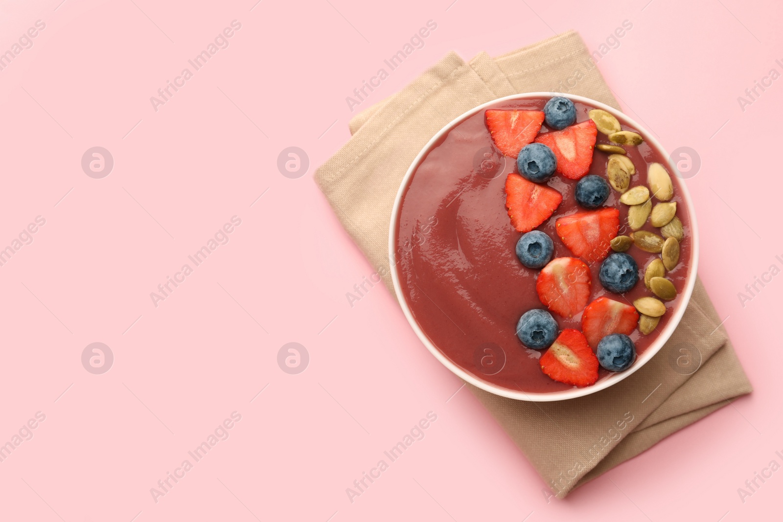 Photo of Bowl of delicious smoothie with fresh blueberries, strawberries and pumpkin seeds on pale pink background, top view. Space for text