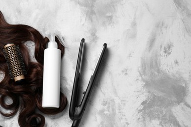 Photo of Spray bottle with thermal protection, lock of brown hair, modern straightener and hairbrush on grey table, flat lay. Space for text