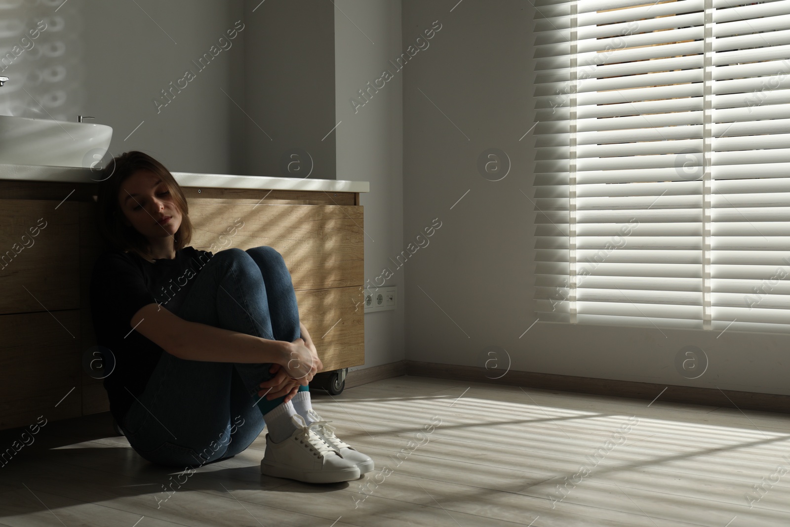 Photo of Sad young woman sitting on floor in bathroom, space for text