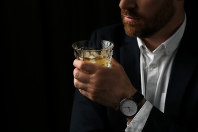 Photo of Man holding glass of whiskey with ice cubes on black background, closeup. Space for text