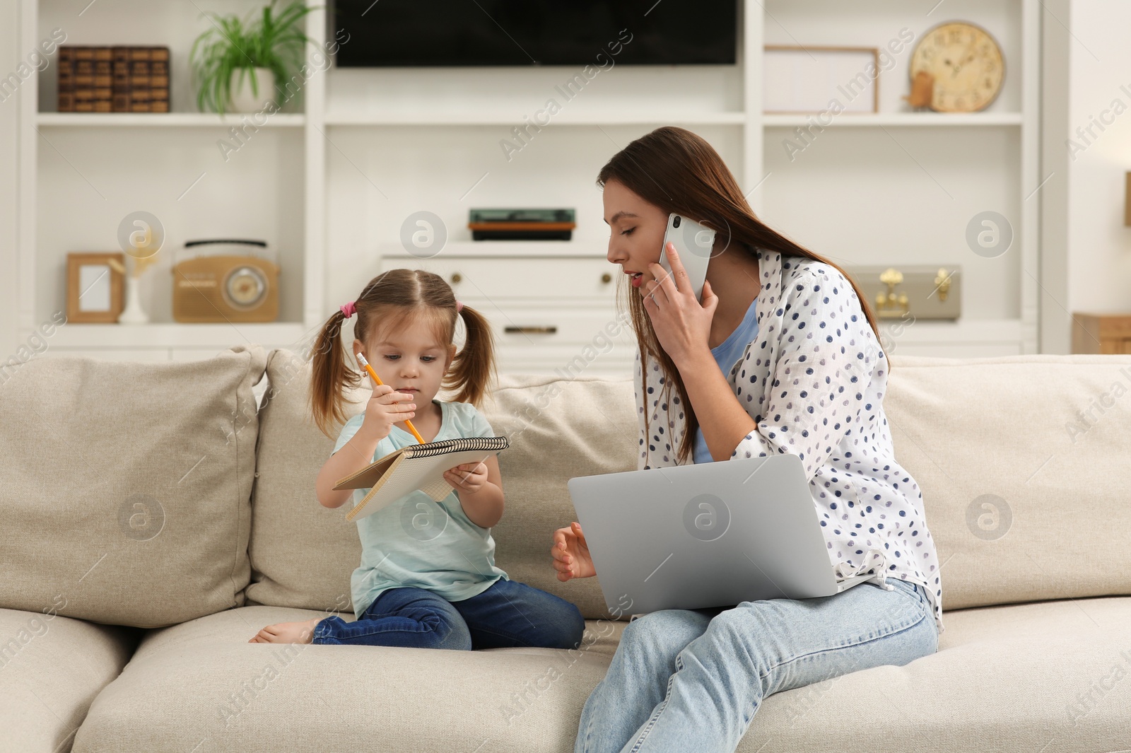 Photo of Working remotely at home. Mother watching over her daughter on sofa in living room. Woman talking on phone while girl drawing in notebook