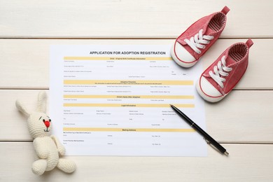 Adoption application, baby shoes, toy bunny and pen on white wooden table, flat lay