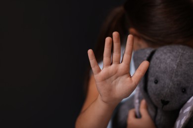 Photo of Child abuse. Little girl with toy bunny doing stop gesture on dark background, selective focus. Space for text