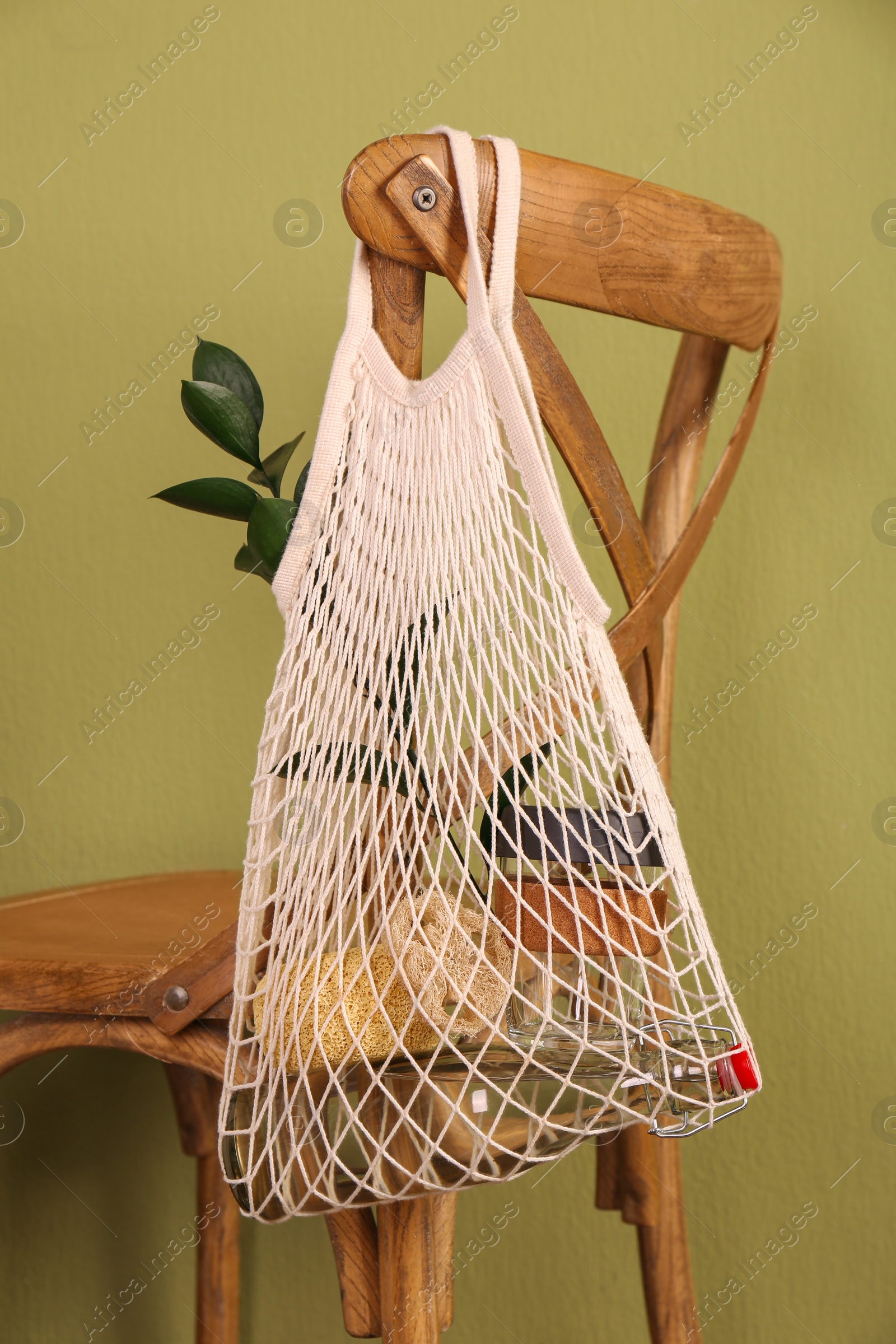 Photo of Conscious consumption. Net bag with eco friendly products hanging on chair near olive wall