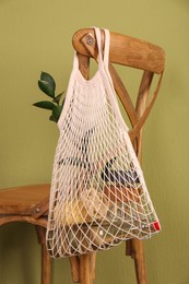 Photo of Conscious consumption. Net bag with eco friendly products hanging on chair near olive wall