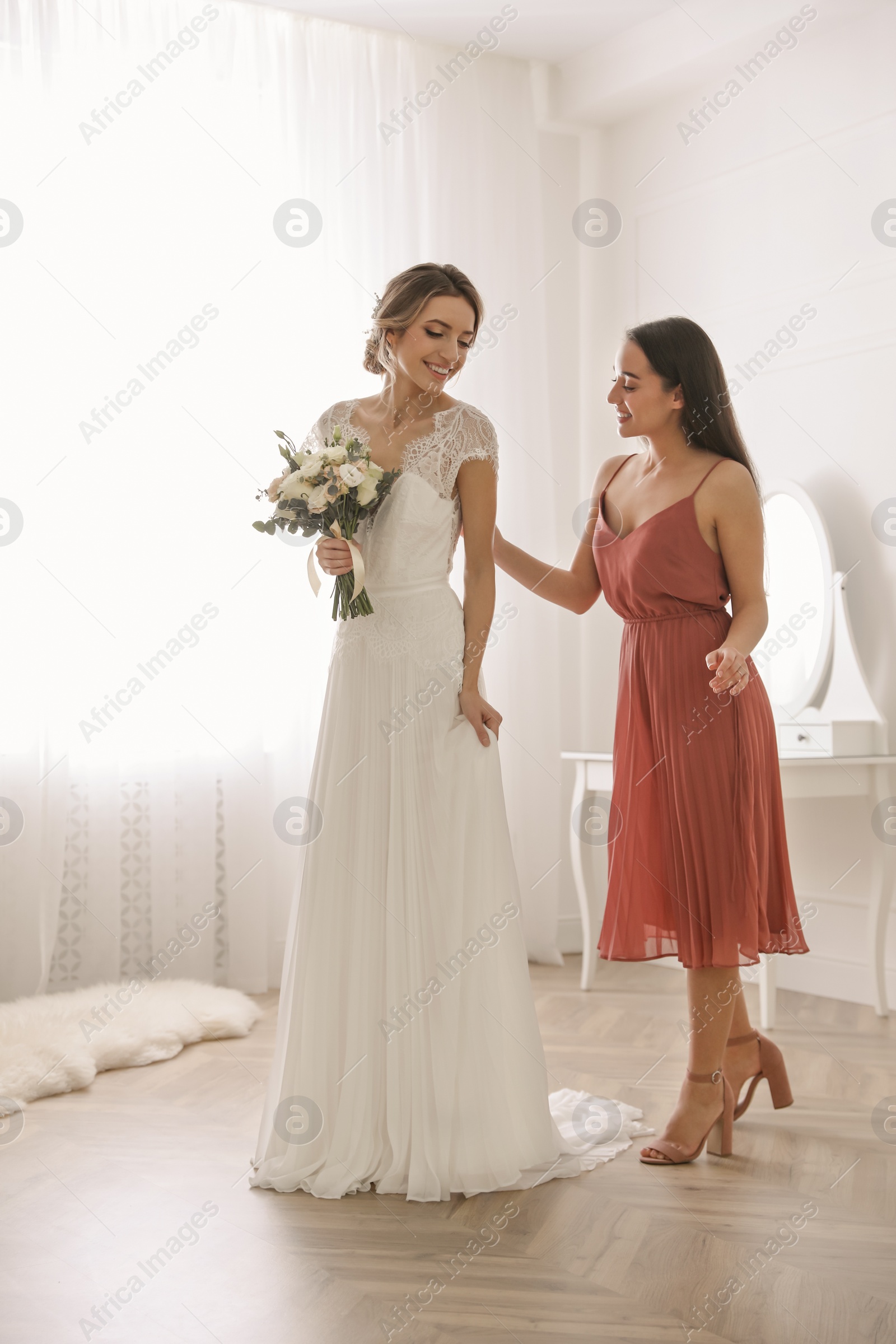 Photo of Young woman helping bride to put on wedding dress in room