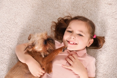 Photo of Portrait of cute girl with funny Brussels Griffon dog lying on carpet, above view. Loyal friends