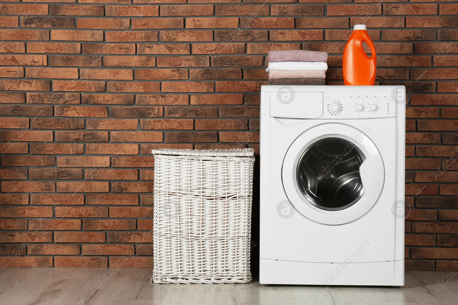 Photo of Modern washing machine with stack of towels, detergent and laundry basket near brick wall