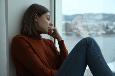Sad young woman sitting on windowsill near window at home