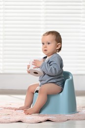 Photo of Little child with toilet paper roll sitting on plastic baby potty indoors