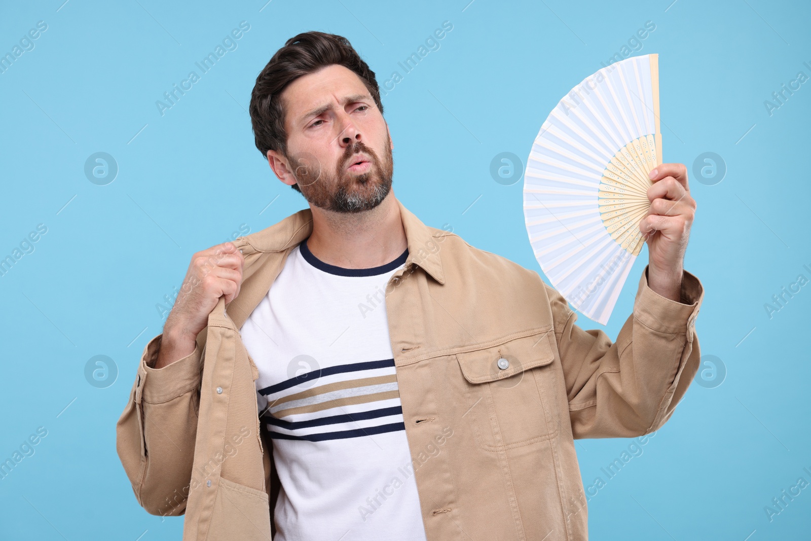 Photo of Unhappy man with hand fan suffering from heat on light blue background