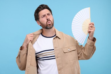 Photo of Unhappy man with hand fan suffering from heat on light blue background