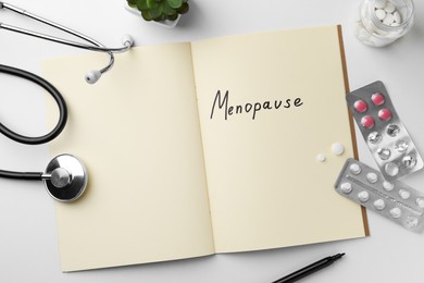 Notebook with word Menopause, pills and stethoscope on white table, flat lay