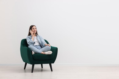 Beautiful woman talking on smartphone while sitting in armchair near white wall indoors, space for text