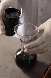 Woman pouring black crude oil from beaker into flask at grey table, closeup