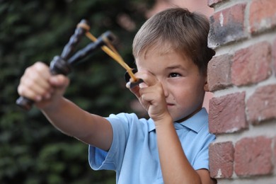 Photo of Little boy playing with slingshot near brick wall outdoors
