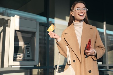 Excited young woman with credit card near cash machine outdoors