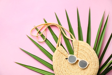 Elegant woman's straw bag with tropical leaf and sunglasses on pink background, top view